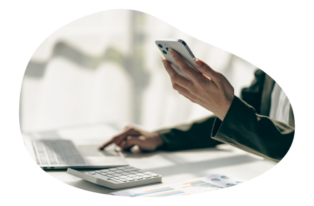 A small business owner using smartphone and laptop with calculator and charts on desk.